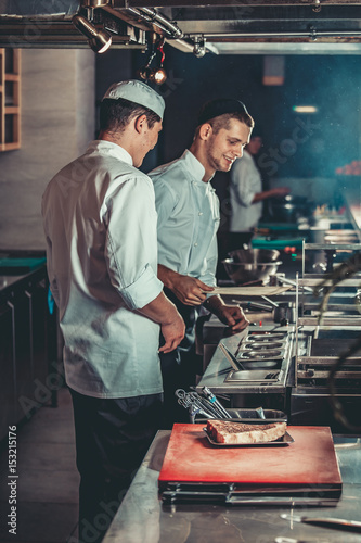 Food concept. Young handsome chefs in white uniform kindle coals and monitor temperature degree for roasting meat in interior of restaurant kitchen. Preparing traditional beef steak on barbecue oven.