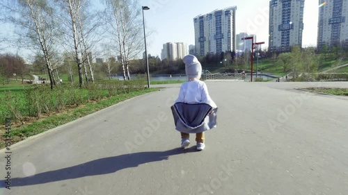 Active child in a hat walks in the park in good weather, making unsteady steps. Little naughty boy runs away, wants to take off his jacket. In the background, high houses, green trees and a pond