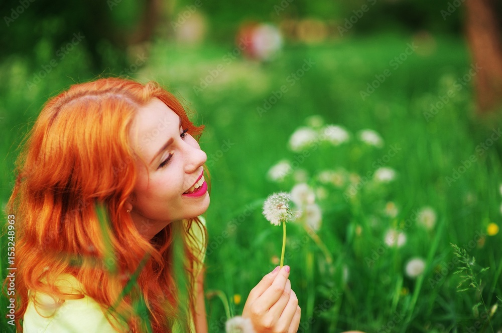 portrait of a beautiful young laughing woman with red hair sitting on meadow with dandelion in hand, blurred background, closeup.