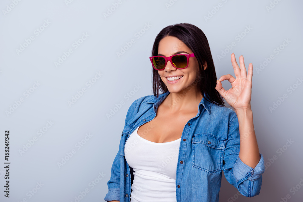 Portrait of a playful young latin girl with in a casual clothes and sunglasses, standing on the blue background and showing ok sign Portrait of a playful young latin girl with in a casual clothes and sunglasses, standing on the blue background and showing ok sign