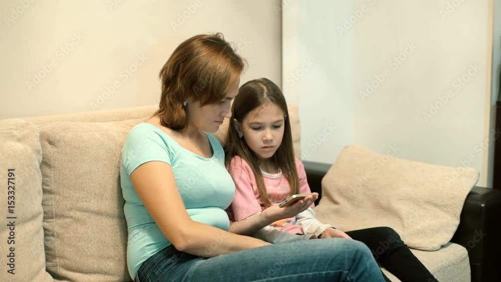 Mom and daughter sitting on sofa watching video on smartphone Mom and daughter sitting on sofa watching video on smartphone