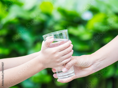 Woman hand giving glass of fresh water to child on the green grass background