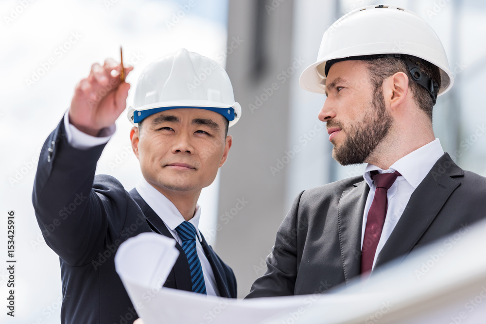 Professional architects in hardhats working with blueprint outside modern building Professional architects in hardhats working with blueprint outside modern building