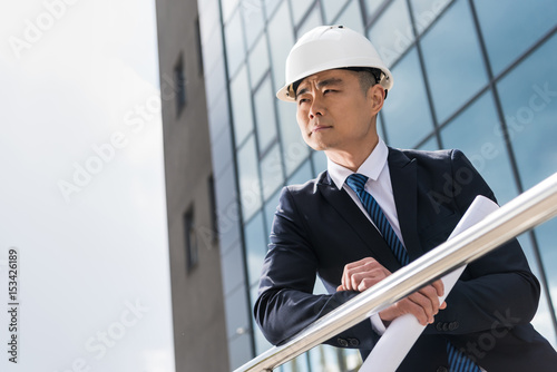 portrait of pensive professional architect in hard hat holding blueprint