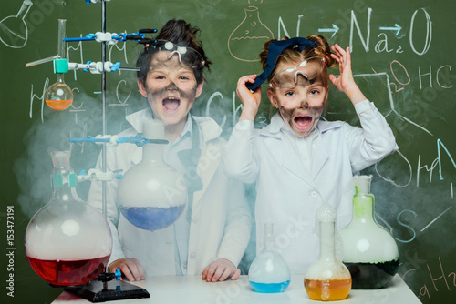 little kids in white coats with chalkboard behind in science laboratory