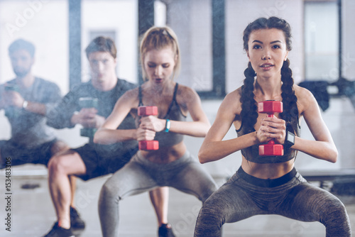 group of athletic young people in sportswear with dumbbells squatting and exercising at the gym