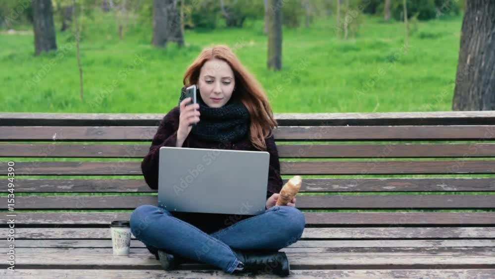 Young woman in scarf working on her laptop and talking on phone in the park Young woman in scarf working on her laptop and talking on phone in the park