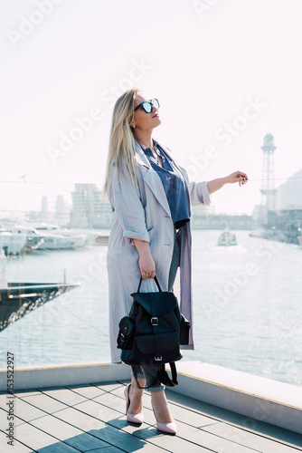 Business woman wearing casual clothes and sunglasses is standing on a high heels at the city view point. Woman is looking aside while standing with the leather backpack in her hand.
