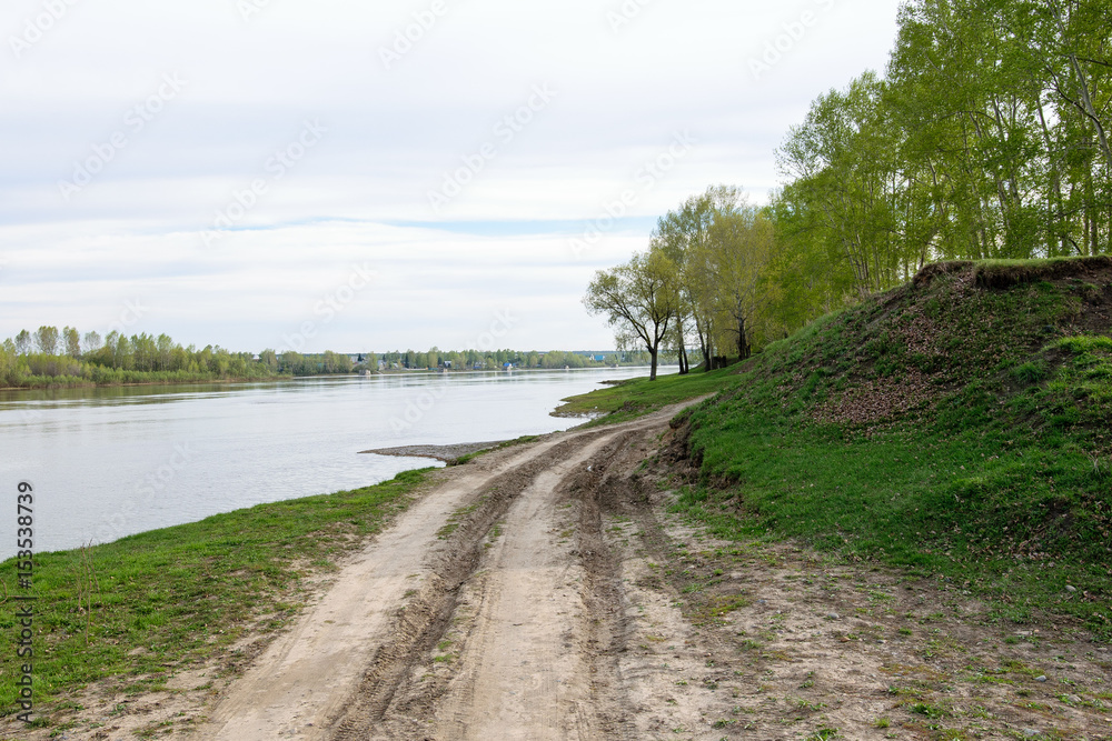 A dirt road on the coast of a calm river in the spring.
