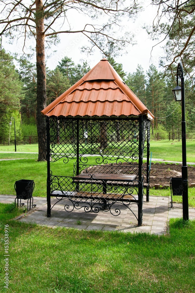 Black gazebo of wrought iron with a pattern and brown tiles in the park on the lawn. Black gazebo of wrought iron with a pattern and brown tiles in the park on the lawn.