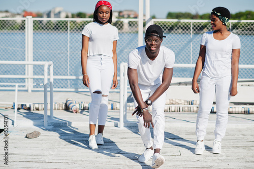 Three stylish african american friends, wear on white clothes at pier on beach. Street fashion of young black people. Black man with two african girls.