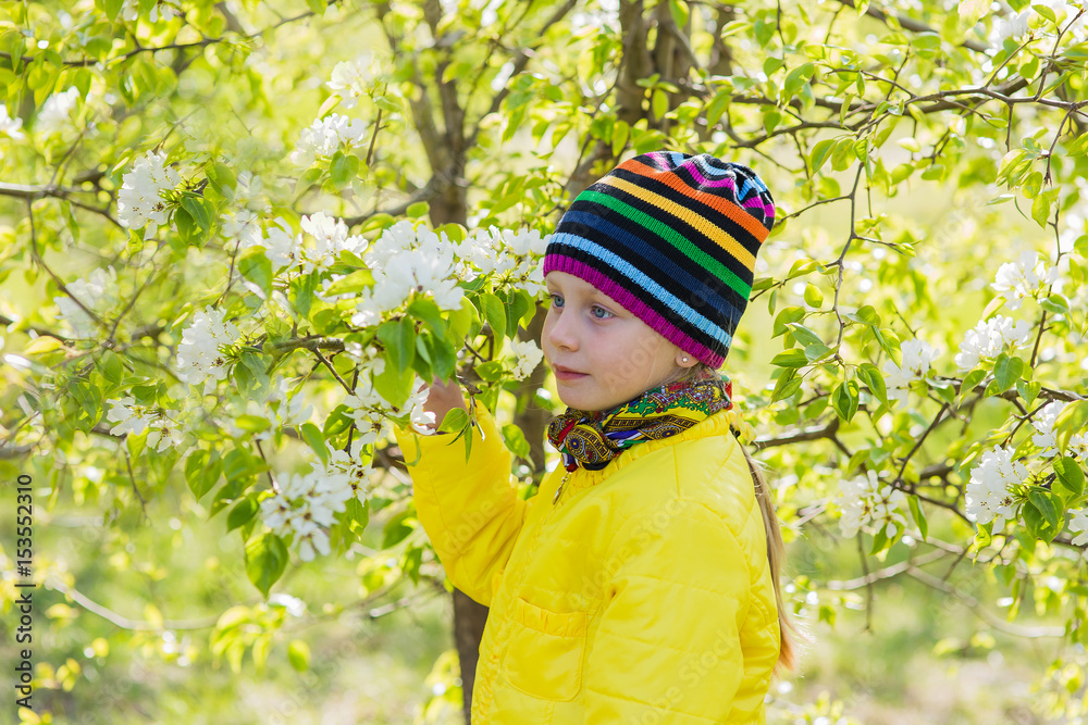 Lovely beautiful girl in a colorful hat and yellow jacket against a blooming apple tree in the spring.