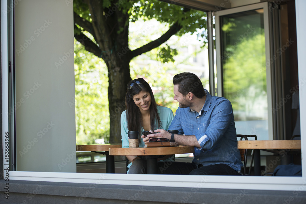 Happy couple looking at mobile phone while sitting in cafe seen through window Happy couple looking at mobile phone while sitting in cafe seen through window