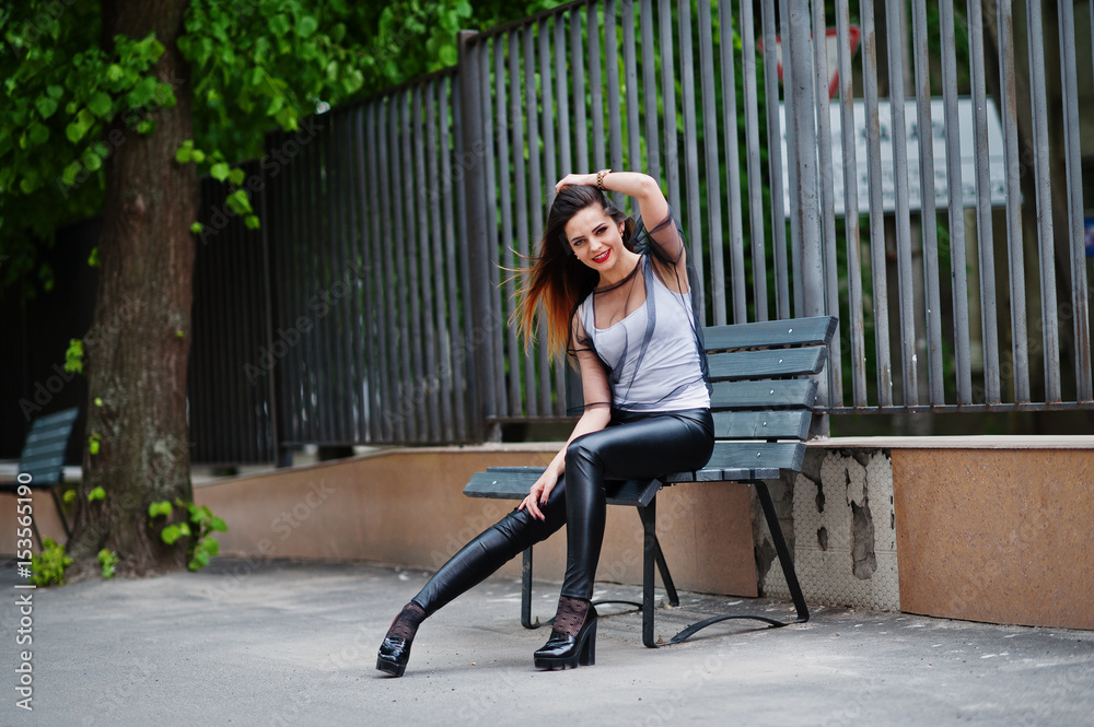 Fashionable woman look at white shirt, black transparent clothes, leather pants, posing at street sitting on bench against iron fence. Concept of fashion girl.