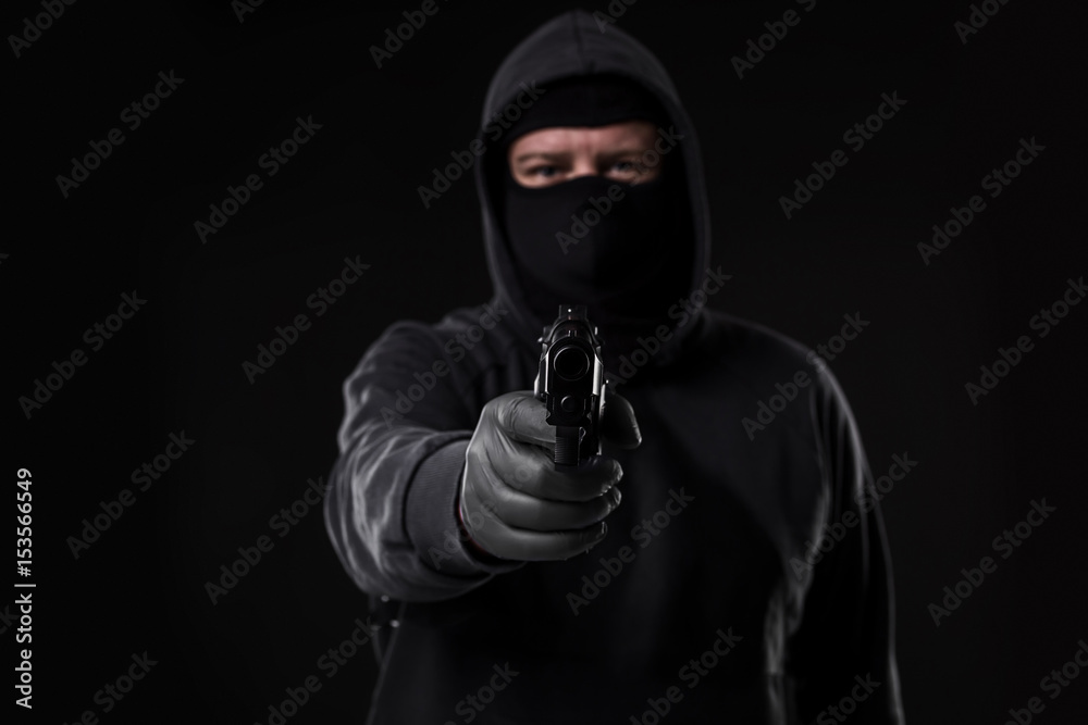 Masked robber with gun aiming into the camera against a black background Masked robber with gun aiming into the camera against a black background
