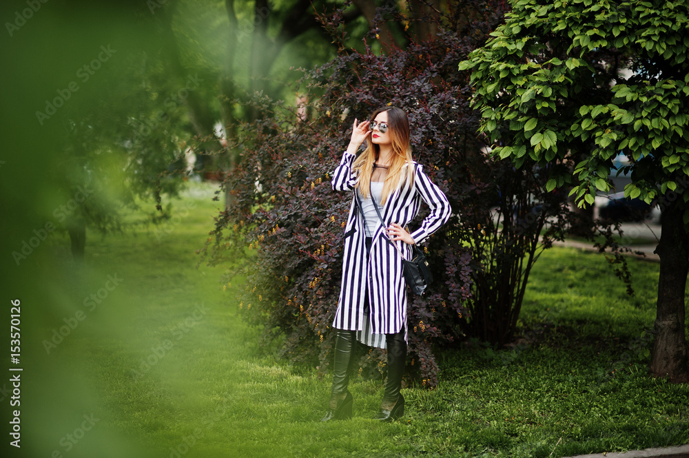 Fashionable woman look with black and white striped suit jacket, leather pants and sunglasses posing against bushes at street. Concept of fashion girl.