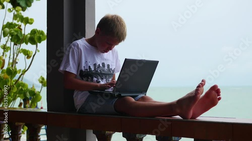 young guy playing on a laptop, sitting on the terrace. Near the sea and the beach