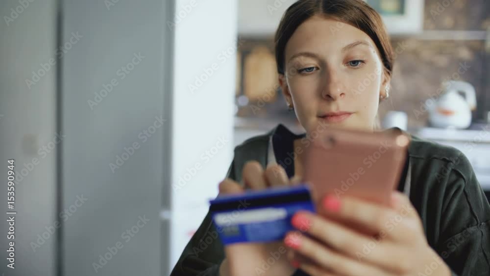 Closeup dolly shot of beautiful young woman have online banking and shopping using smartphone and credit card in the kitchen at home Closeup dolly shot of beautiful young woman have online banking and shopping using smartphone and credit card in the kitchen at home