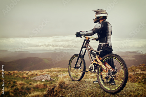 Fototapeta Naklejka Na Ścianę i Meble -  The rider in the full-face helmet and full protective equipment on the mtb bike stands on a rock against the background of a ridge and low clouds