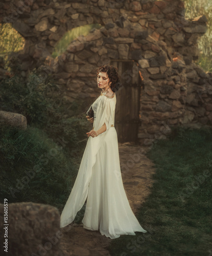 Young vintage woman walk near castle. adult beautiful girl in Greek white medieval historical dress holding book in hand. princess stands, old stone courtyard path way. warm colors back rear view