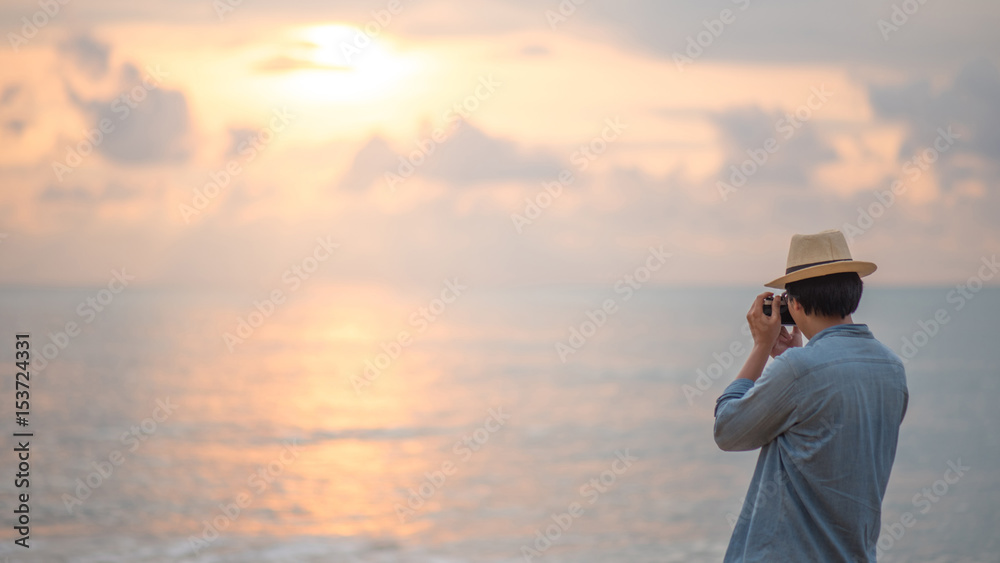 Young Asian man traveler and photographer with jean shirt and hat taking photos of beautiful sunset at tropical beach island, background for summer holiday and vacation travel concepts