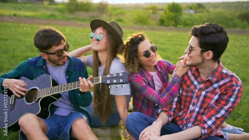 Four friends plays guitar and sings songs outdoors at sunset. They very happy and cheerful