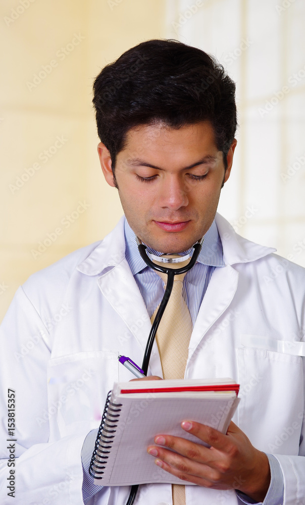 Handsome doctor with an Stethoscope holding from his neck, writing on his notebook notes in office background Handsome doctor with an Stethoscope holding from his neck, writing on his notebook notes in office background