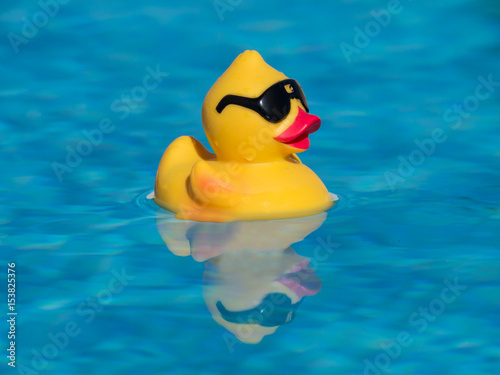 Fotografia Yellow rubber duck with black sunglasses floating on a beautiful blue swimming pool