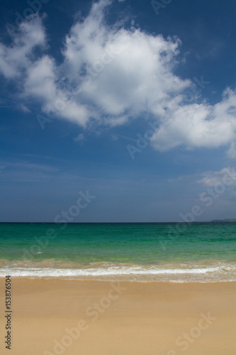 A deserted sandy beach and an emerald green ocean under a bright blue sky with white, fluffy clouds on a deserted paradise island.