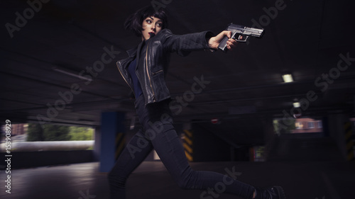 Beautiful brunette sexy spy agent (killer or police) woman in leather jacket and jeans with a gun in her hand running after someone, to catch him on parking