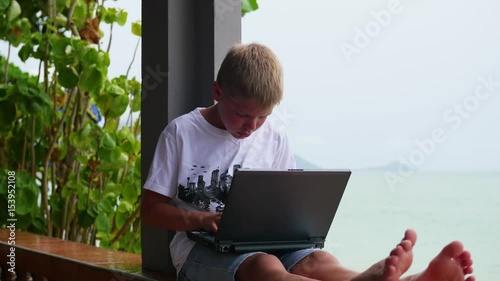 young guy playing on a laptop, sitting on the terrace. Near the sea and the beach