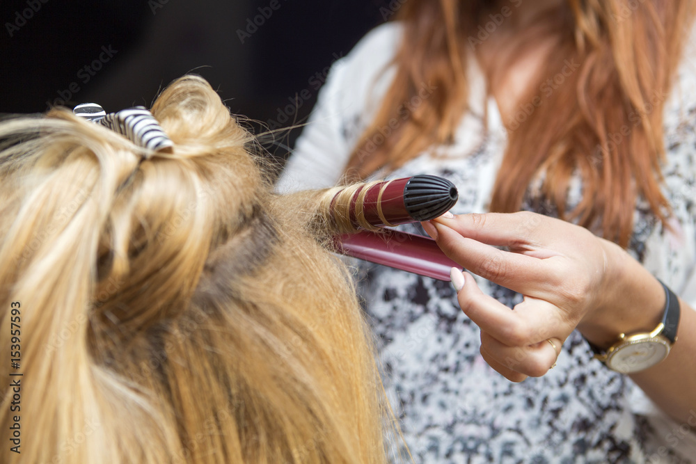 Fototapeta premium Brunette red hair hairdresser artist making curly hairstyle to blonde bride woman on her wedding day with iron. Studio interior, close up of a hands in process