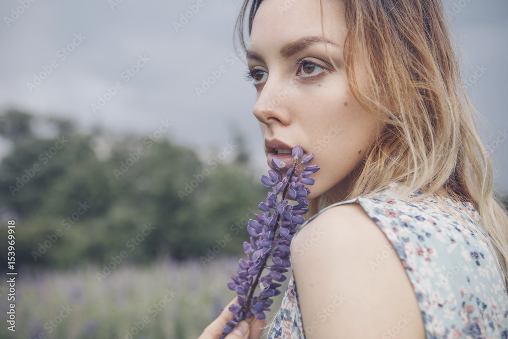 Fototapeta premium Beautiful brunette fit slim fragile woman with clear flawless skin and waved hair in sensual summer dress in the middle of meadow lupins field on a sunset, looking thoughtful and sad