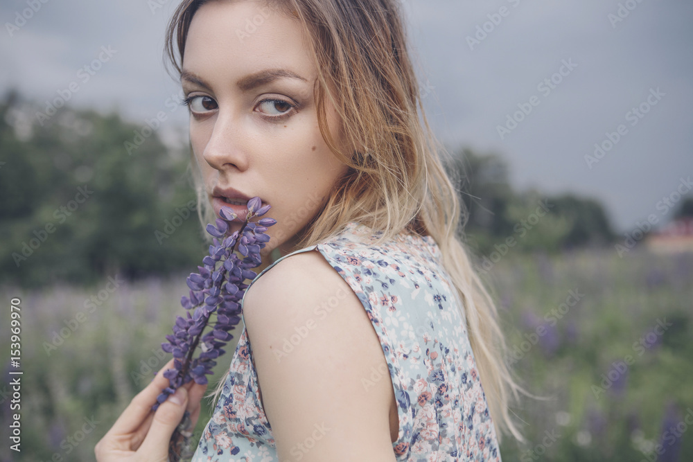 Naklejka premium Beautiful brunette fit slim fragile woman with clear flawless skin and waved hair in sensual summer dress in the middle of meadow lupins field on a sunset, looking thoughtful and sad