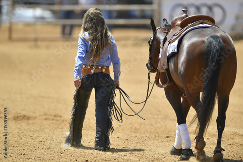 The rear view of a rider in cowboy chaps and boots leads the horse out from the sand field