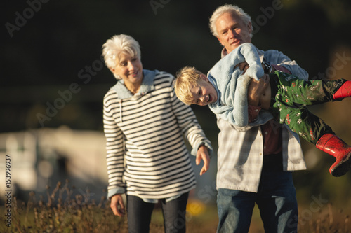 Small boy laughs as his grandfather playfully hefts him up horizontally and his grandmother smiles as she watches while they walk together outside. photo