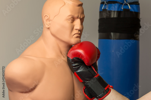 Red boxing gloves punching an uppercut on the chin of a dummy mannequin doll on a gym with a blue punching bag on the background