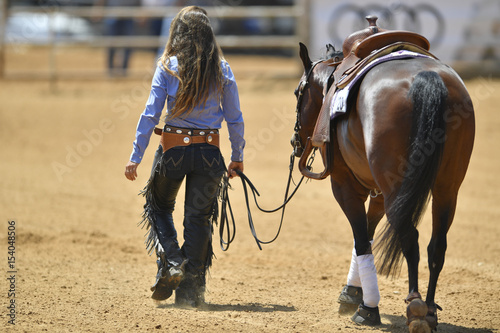 The rear view of a rider in cowboy chaps and boots leads the horse out from the sand field