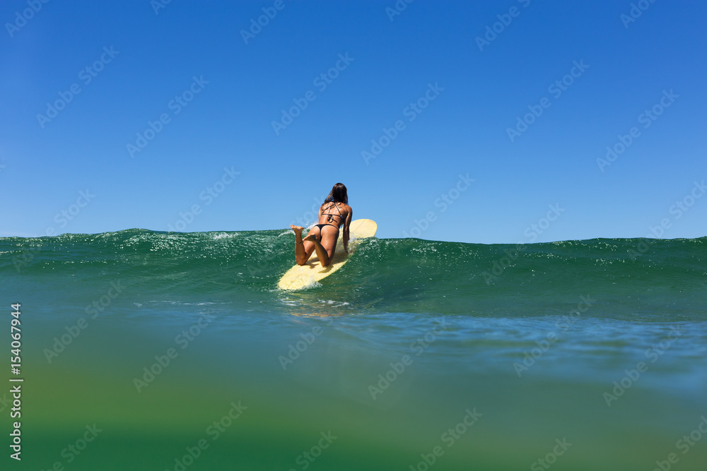 A beautiful young surfer girl in a bikini paddles over a clear ocean wave beneath a blue summer sky.