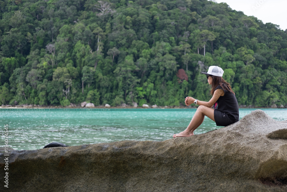 woman sitting on rock. front of her have blue sea and green forest on mountain. this image for people,nature and landscape concept