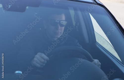 Мужчина в солнцезащитных  очках сидит в тонированной машине и держится руками за руль. Вид с лобового окна автомобиля

