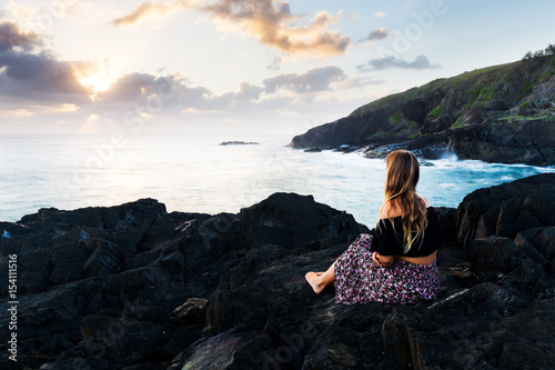 A beautiful bohemian-styled girl sits on a rocky headland and watches the morning sun burst above the clouds during a magnificent sunrise in Australia.