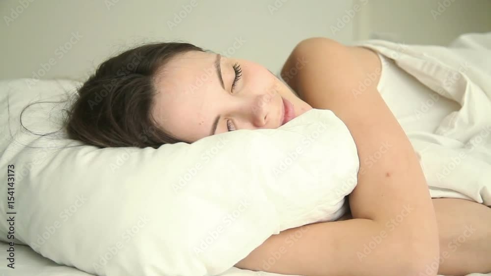 Young attractive happy girl sleeping soundly on bed in bedroom at home or hotel, cuddling white comfy pillow, enjoying good dream, lying with her eyes closed on comfortable mattress in relaxed pose Young attractive happy girl sleeping soundly on bed in bedroom at home or hotel, cuddling white comfy pillow, enjoying good dream, lying with her eyes closed on comfortable mattress in relaxed pose