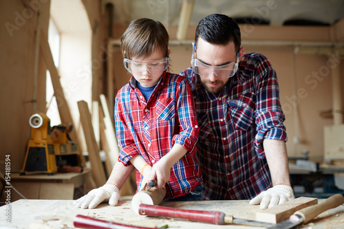 Youthful kid with chisel carving wooden workpiece with his father near by
