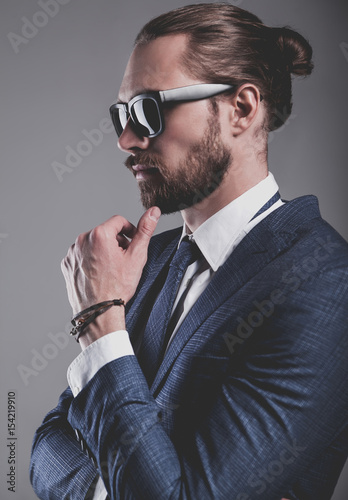 portrait of handsome fashion businessman  model dressed in elegant blue suit with sunglasses.posing on gray background in studio. Touching his beard photo