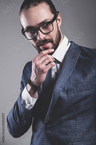 portrait of handsome fashion businessman  model dressed in elegant blue suit with glasses.posing on gray background in studio. Touching his beard photo