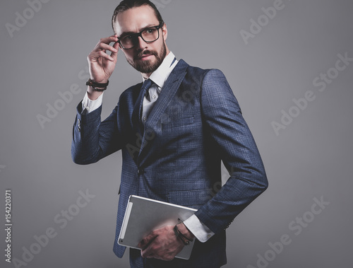 portrait of handsome fashion businessman  model dressed in elegant blue suit with glasses.posing on gray background in studio. Using touchpad photo