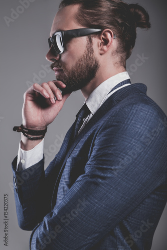 portrait of handsome fashion businessman  model dressed in elegant blue suit with sunglasses.posing on gray background in studio. Touching his beard photo