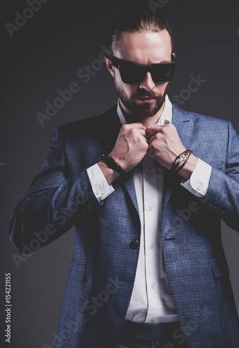 portrait of handsome fashion stylish hipster businessman model dressed in elegant blue suit posing on gray background in studio. Buttoning his jacket