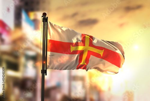 Guernsey Flag Against City Blurred Background At Sunrise Backlight